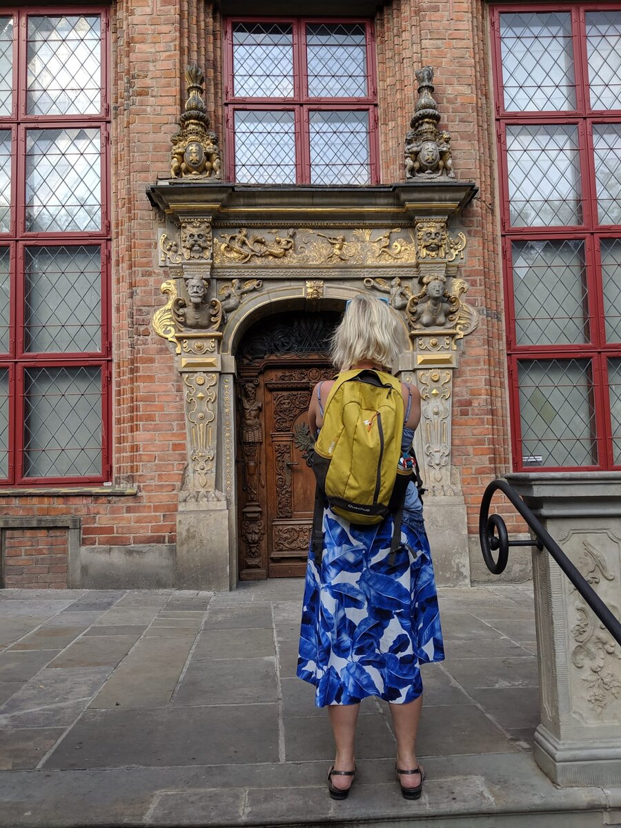 A blonde woman seen from behind, wearing a blue-and-white leaf-pattern skirt and yellow backpack, standing before an ornate gilded Renaissance doorway of a Gdansk merchant house.