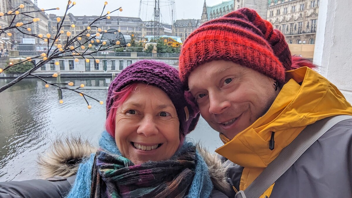 Kirsten and Rob in a close selfie on a walkway over the Alster in Hamburg, winter. Kirsten in a purple knit hat and fur-trimmed coat, pink hair peeking out. Rob in a red-orange beanie and yellow rain jacket, smiling gently. The Rathaus spire and decorative winter lights on bare branches behind them.