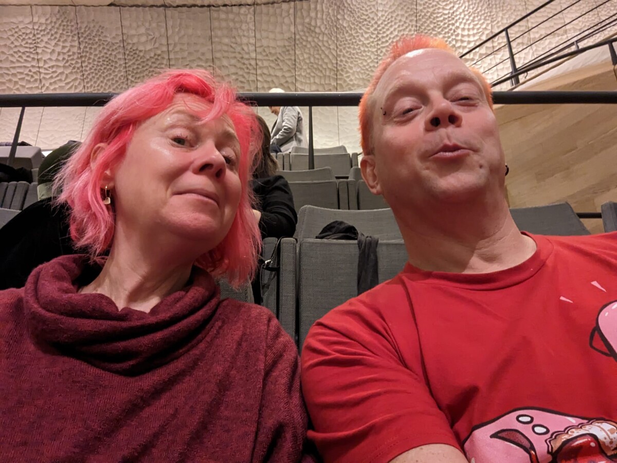 Selfie of Kirsten with pink hair in a burgundy sweater and Rob with pinkish-orange hair in a red cartoon t-shirt, seated in the Elbphilharmonie concert hall with its distinctive white acoustic wall panels behind them. Rob is making a duck-face expression.