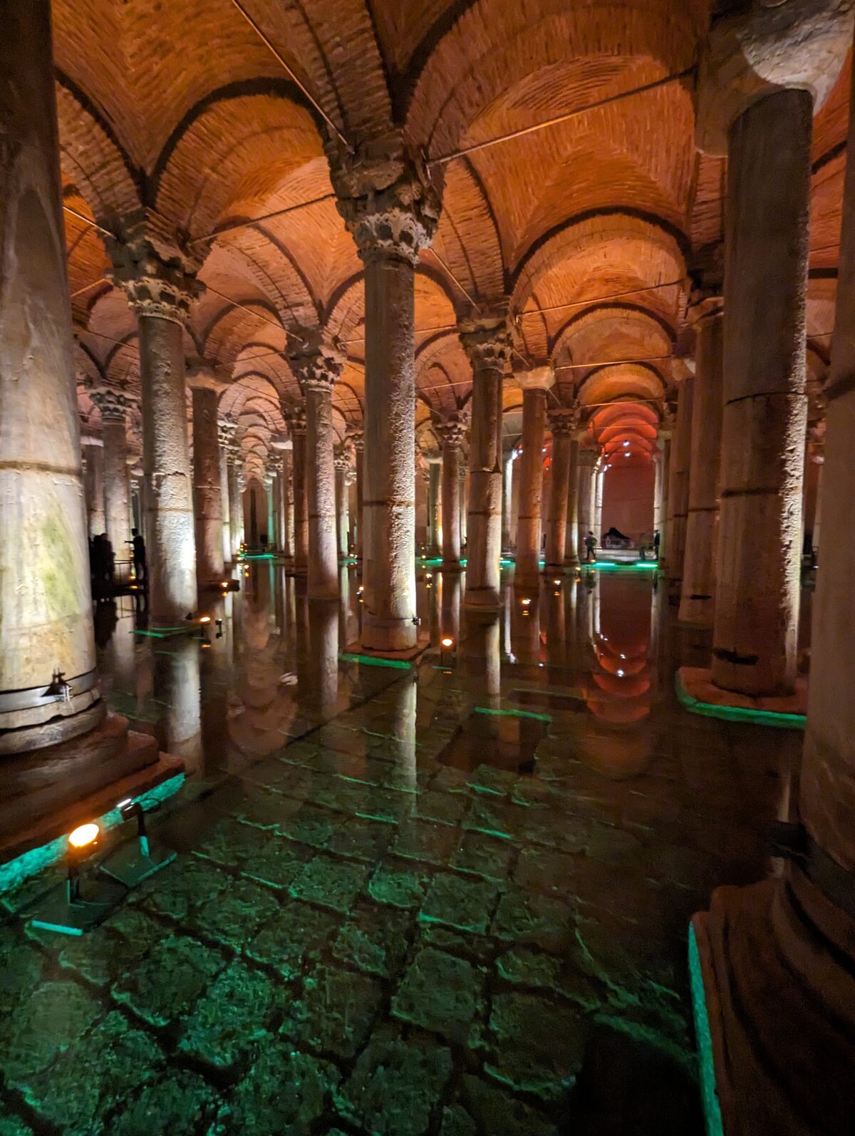 Long colonnade of repeating stone columns stretching into the distance inside the Basilica Cistern, reflected in still green-lit water below, with warm orange uplights on the columns and a dark vaulted brick ceiling above.
