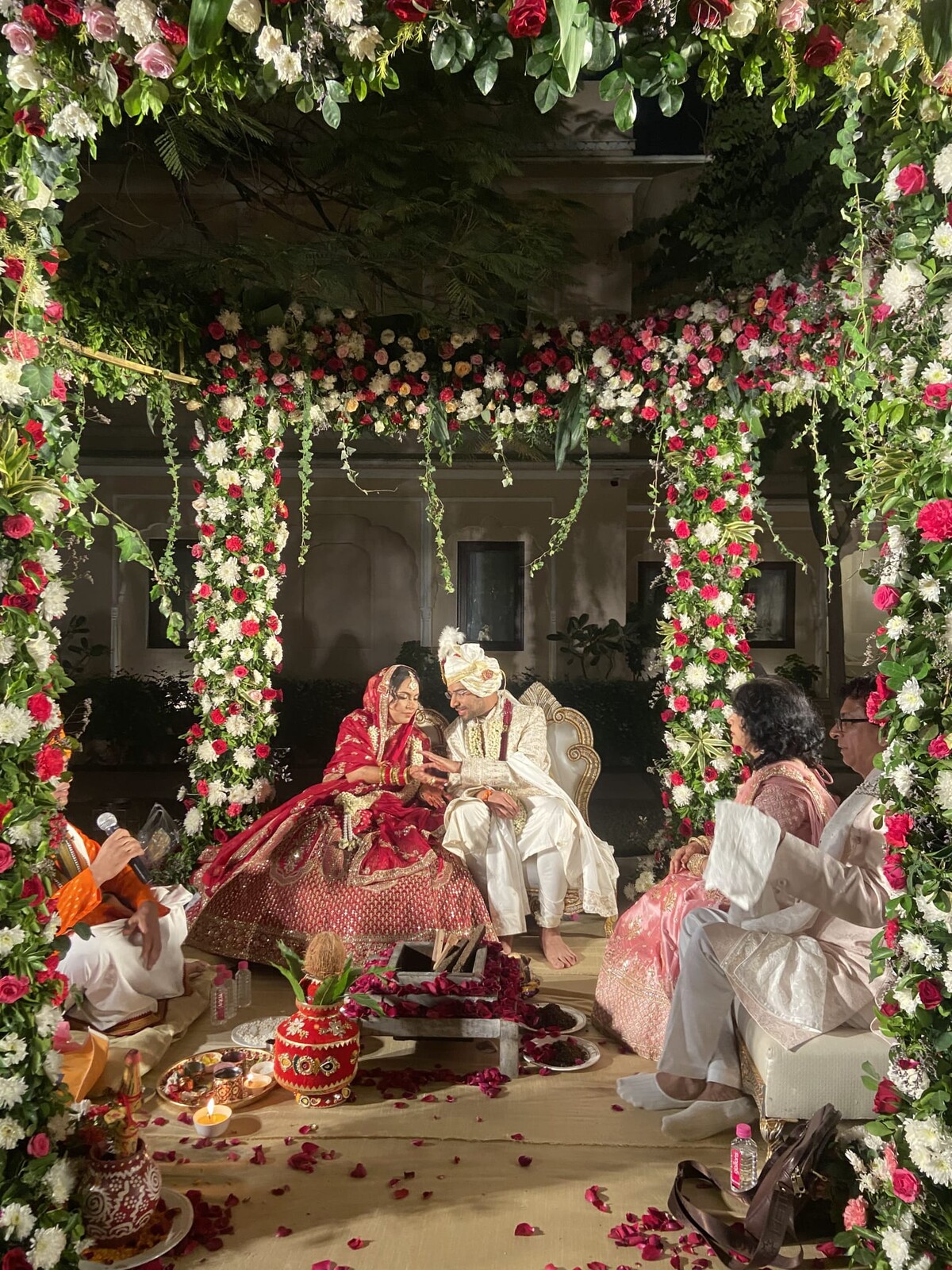 Hindu wedding ceremony under a mandap canopy framed by pillars of red, pink, and white roses. The bride in traditional red and gold and the groom in cream are seated at center with a priest, warm night lighting creating a golden glow.