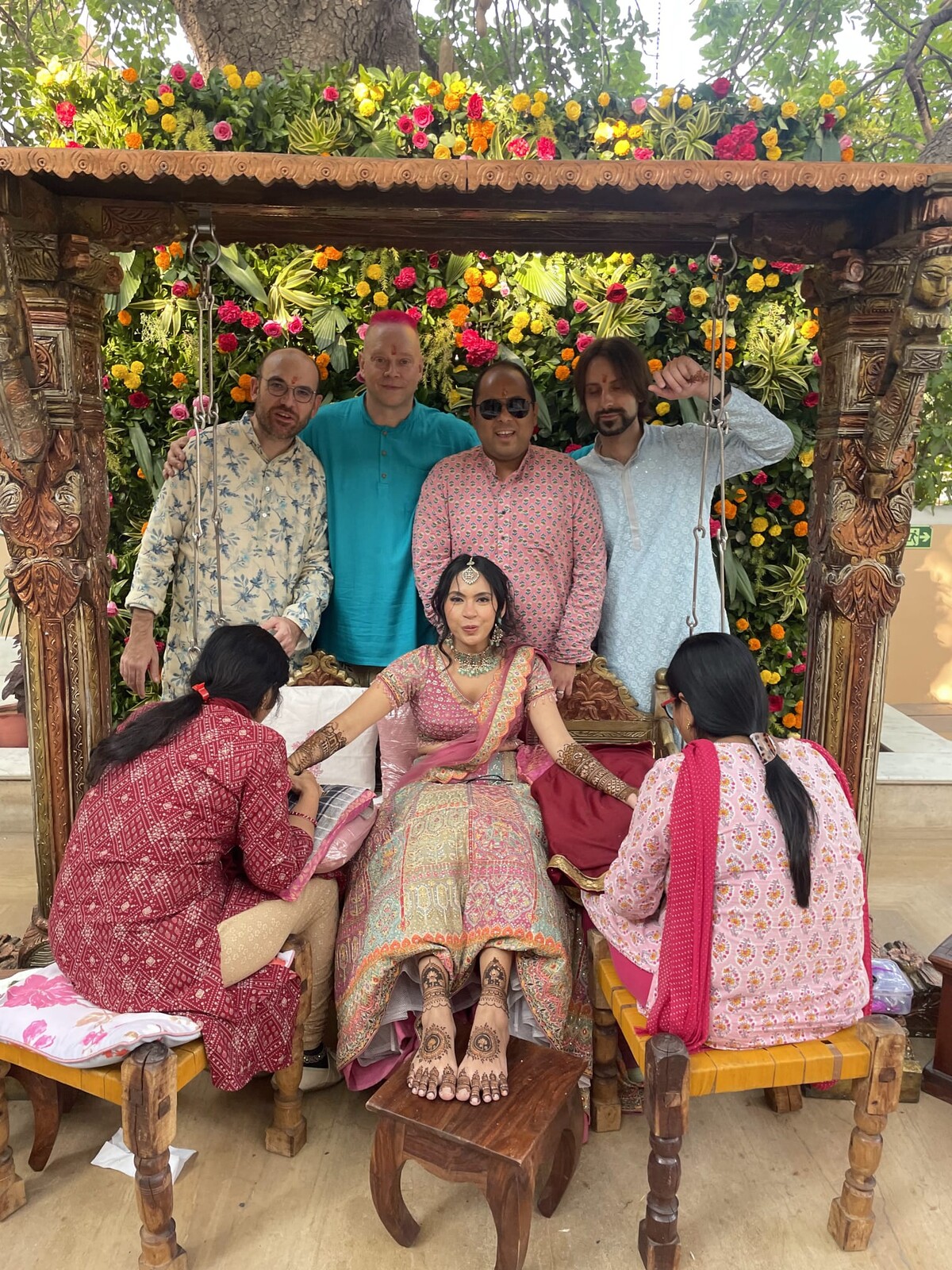 Group portrait framed by an ornately carved wooden swing draped in marigolds and roses. The bride sits center on the swing while four men stand behind her, including Rob in a teal kurta. Two women attend to the bride in the foreground.