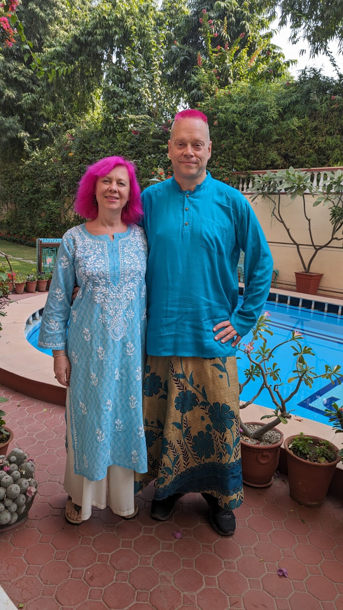 Rob and Kirsten standing by a hotel swimming pool in India. Rob has pink hair and wears a teal kurta with a gold-and-teal floral skirt; Kirsten has pink hair and wears a light blue embroidered salwar kameez. Both look relaxed and happy.