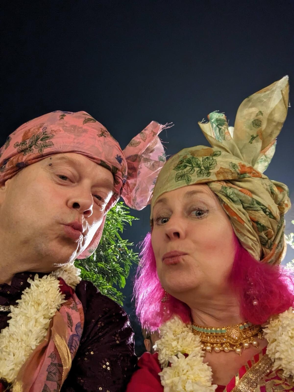 Nighttime selfie of Rob in a pink patterned turban and jasmine garland making a duck face, and Kirsten in an elaborate green-and-orange turban and jasmine garland making a matching expression, both laughing.