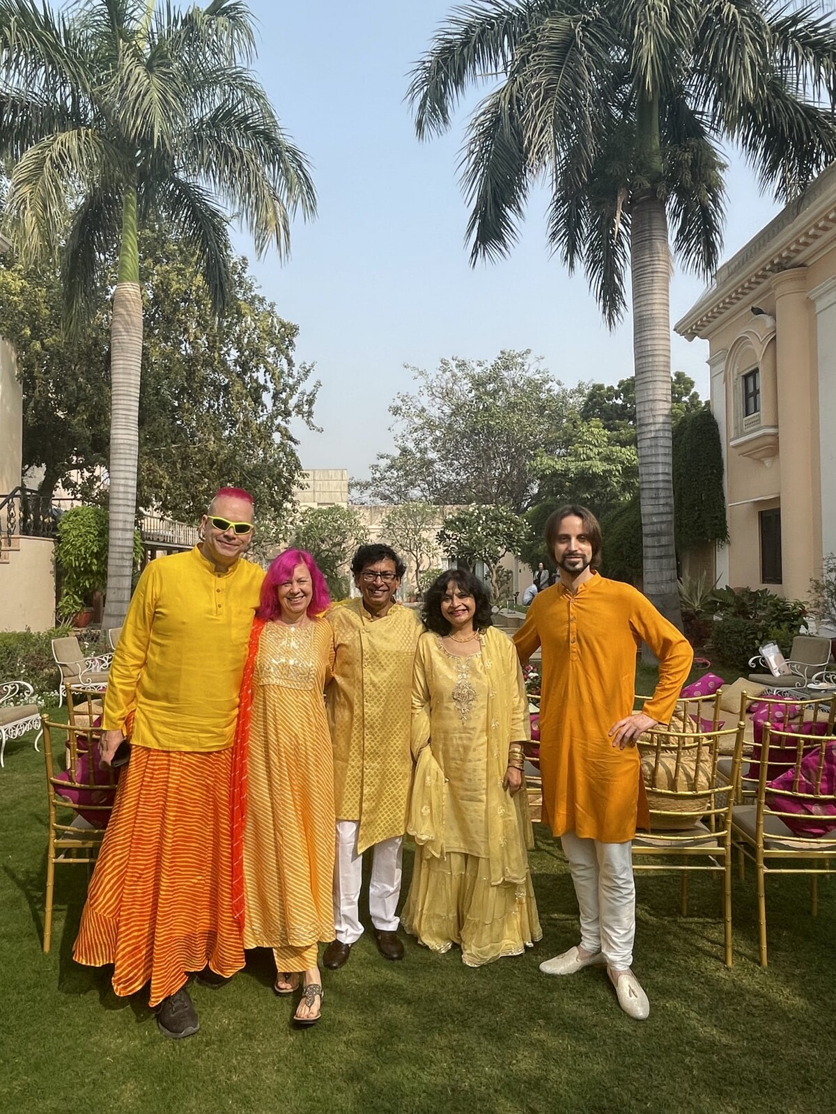 Five people standing on a manicured palace lawn with palm trees, all wearing traditional Indian wedding attire in coordinated yellow-orange-gold colors. Rob on the far left with pink hair, Kirsten beside him, three friends completing the group.