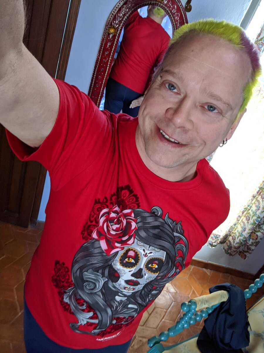 Rob with multicolored yellow-green and pink hair, proudly displaying a red Day of the Dead Catrina skull t-shirt in a mirror selfie, turquoise beaded jewelry visible.