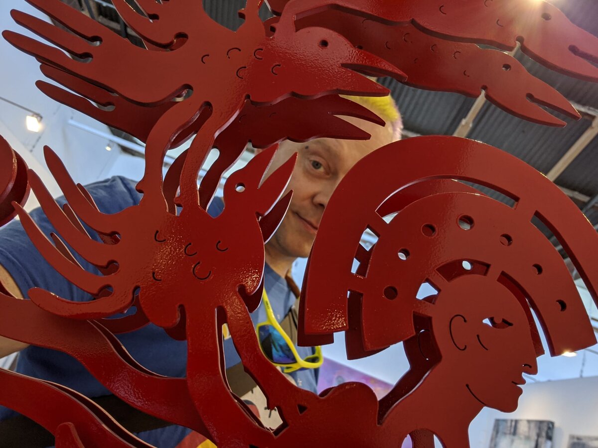 Rob's face peeking through gaps in an intricate red laser-cut metal sculpture with Aztec-inspired figures and headdresses, in a gallery.