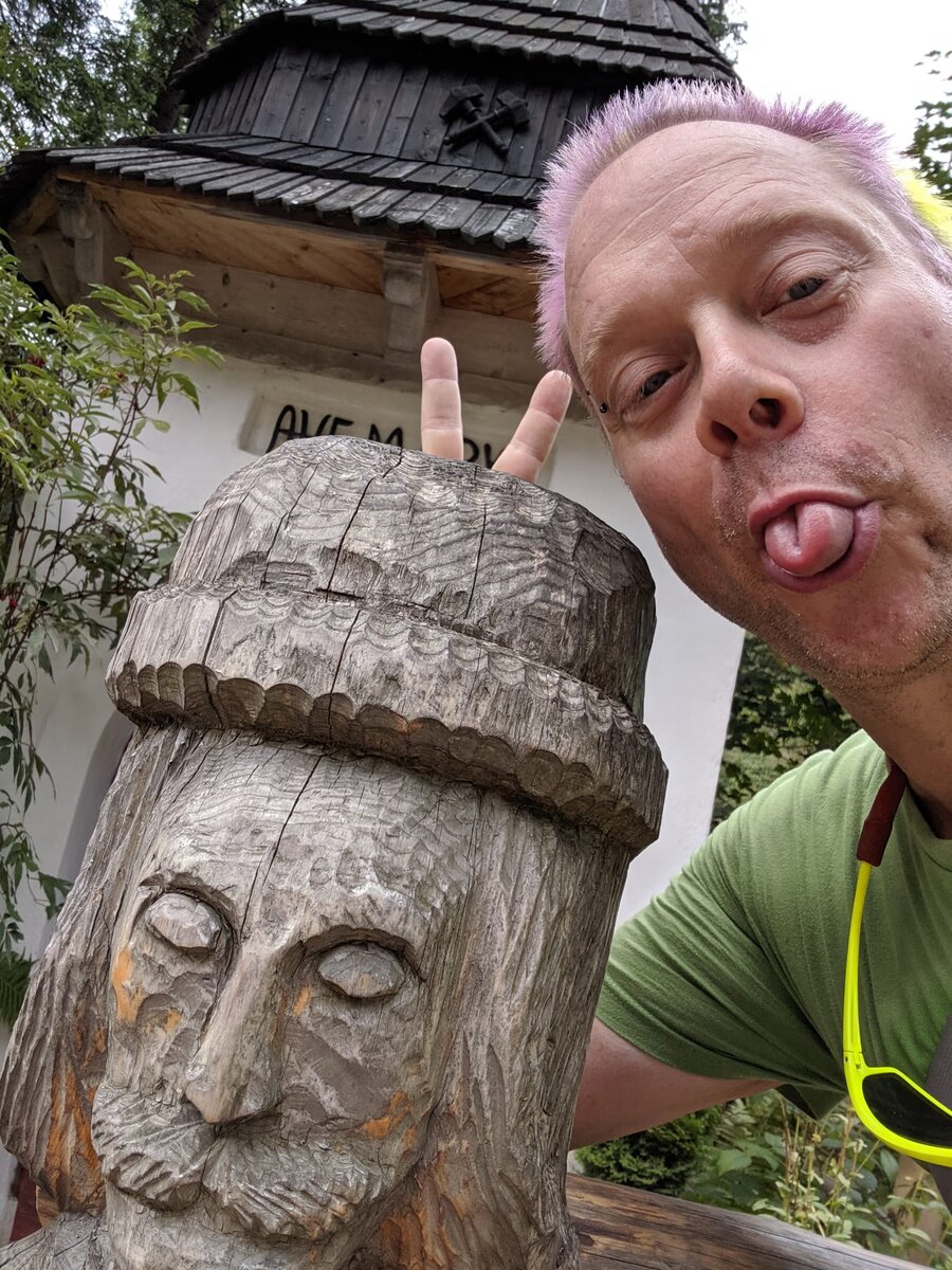 Rob with pink-purple hair, tongue sticking out and hand making a rock-on gesture, posing next to a carved wooden folk figure of a bearded man in a hat, with a wooden bell tower behind them in the Tatra Mountains.