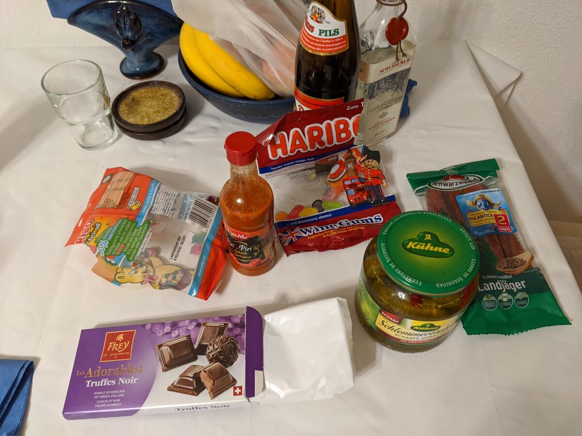 Overhead flat-lay of Rob's holiday provisions: German beer, Haribo gummy bears, Wine Gums, piri-piri hot sauce, pickles, dried sausage, Swiss dark chocolate truffles, and bananas on a white cloth.