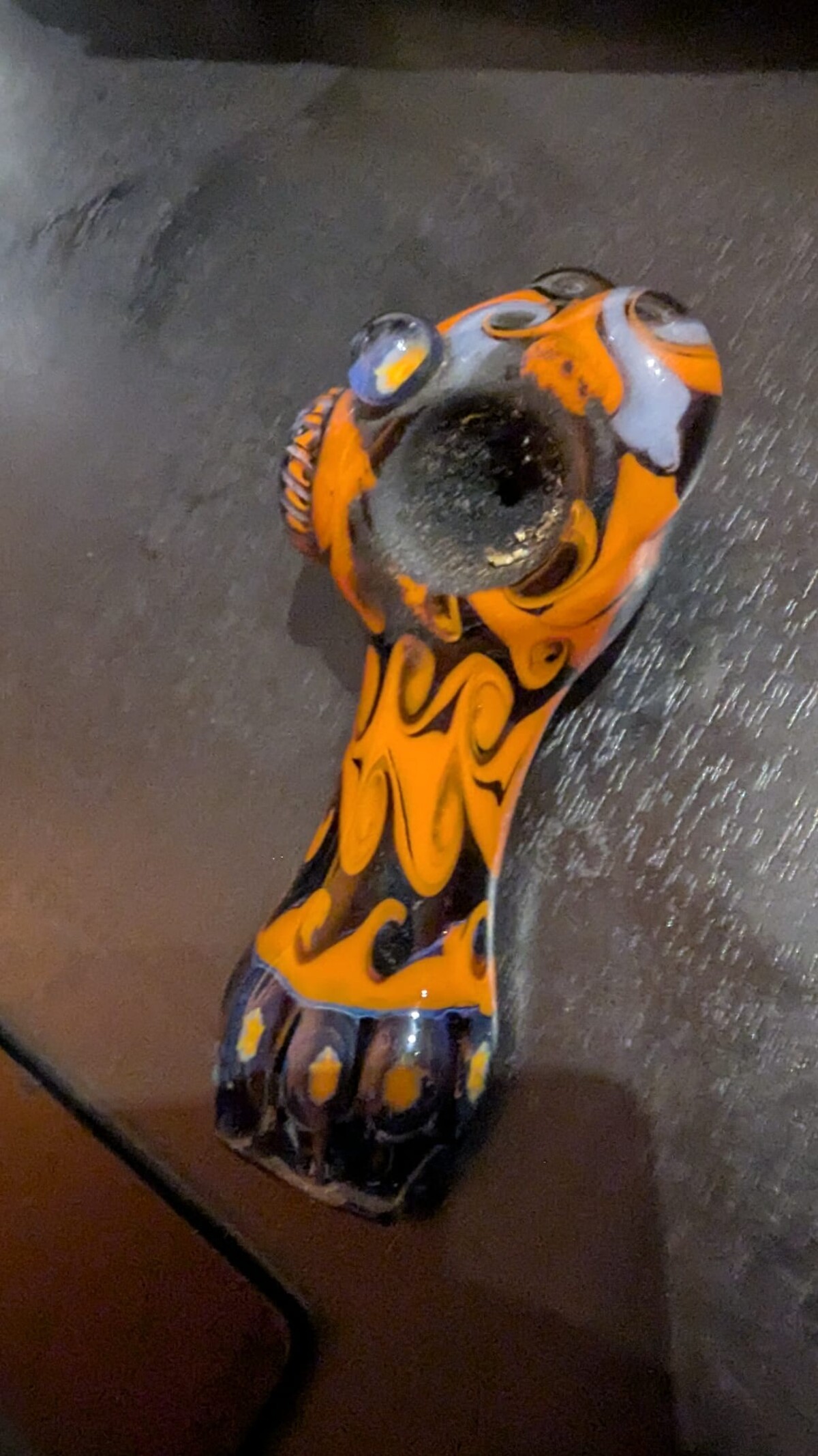 Close-up overhead shot of a hand-blown glass pipe with intricate orange and black swirl patterns, purple and blue accent dots, resting on a dark surface. The bowl is blackened from use.