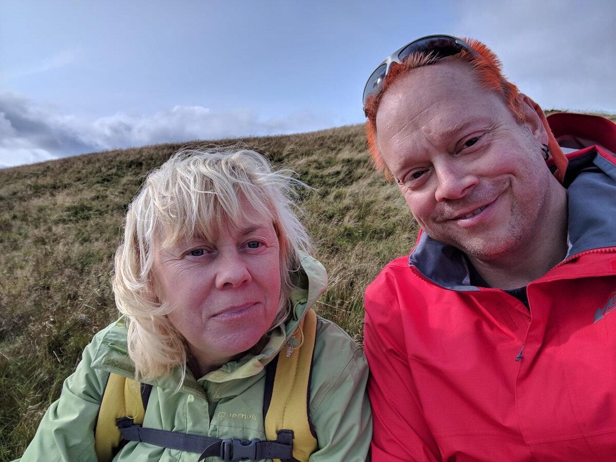 Rob with orange hair in a red rain jacket and Kirsten in a green Quechua jacket, close selfie on a Scottish hillside, windblown hair, overcast sky.