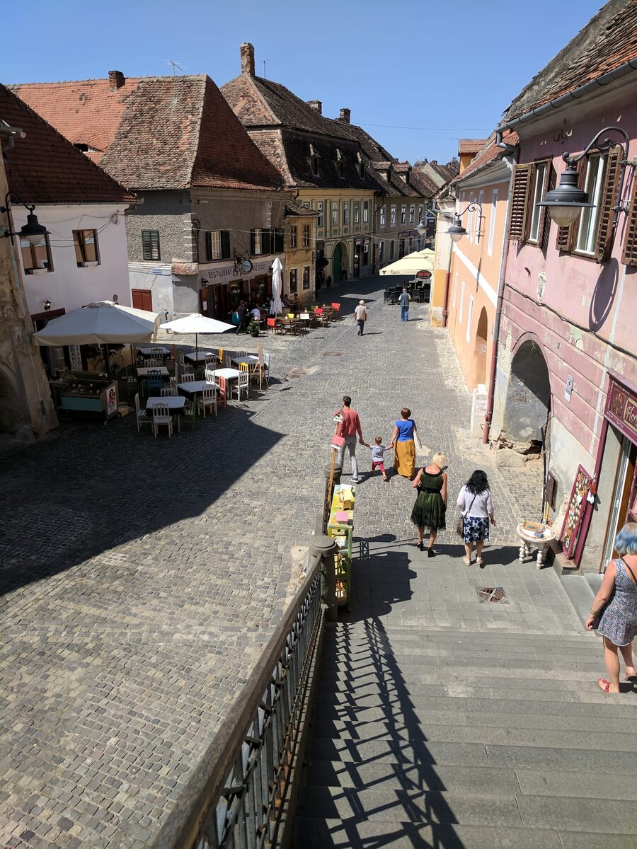 View from stone steps down into a sunlit cobblestoned Transylvanian town square, Saxon-style pastel buildings with steep terracotta roofs, outdoor cafe tables with white umbrellas.