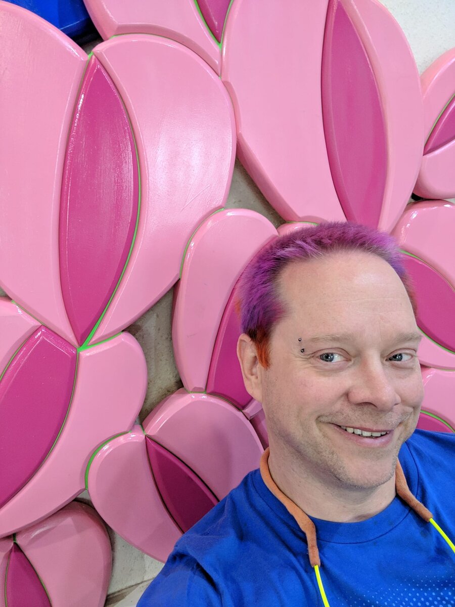 Rob with purple hair grinning broadly against a backdrop of large pink sculptural petals at a public art installation, his hair echoing the pink tones.