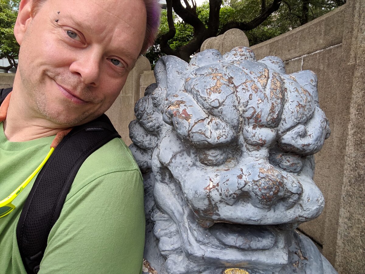 Rob with purple hair and a bridge piercing taking a close selfie next to a weathered blue-grey stone guardian lion statue at a temple.