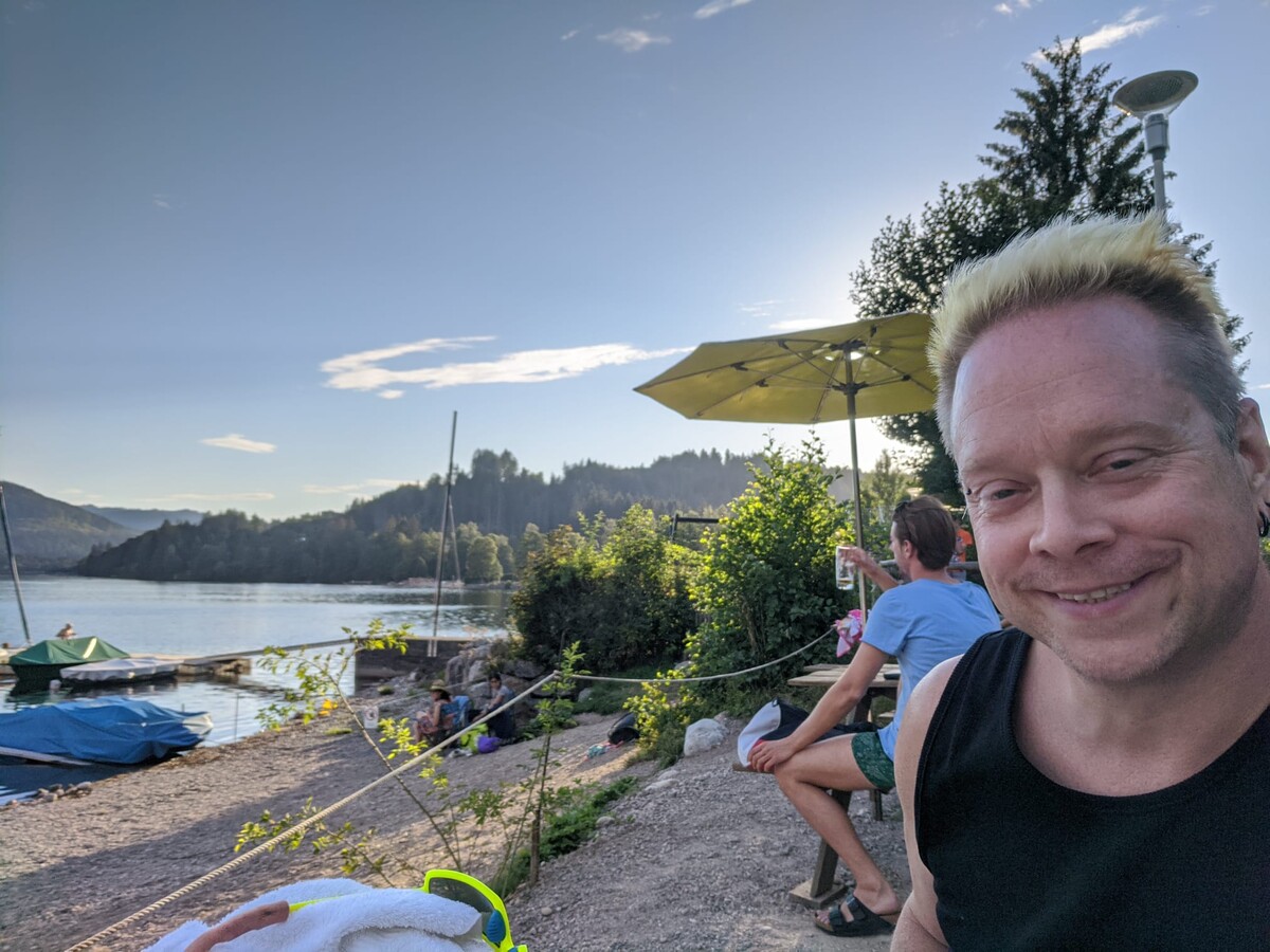 Rob taking a golden hour selfie at Titisee lake in the Black Forest, platinum blonde hair spiked upward, wearing a black tank top. Forested hills, a yellow parasol, and the lake behind him.