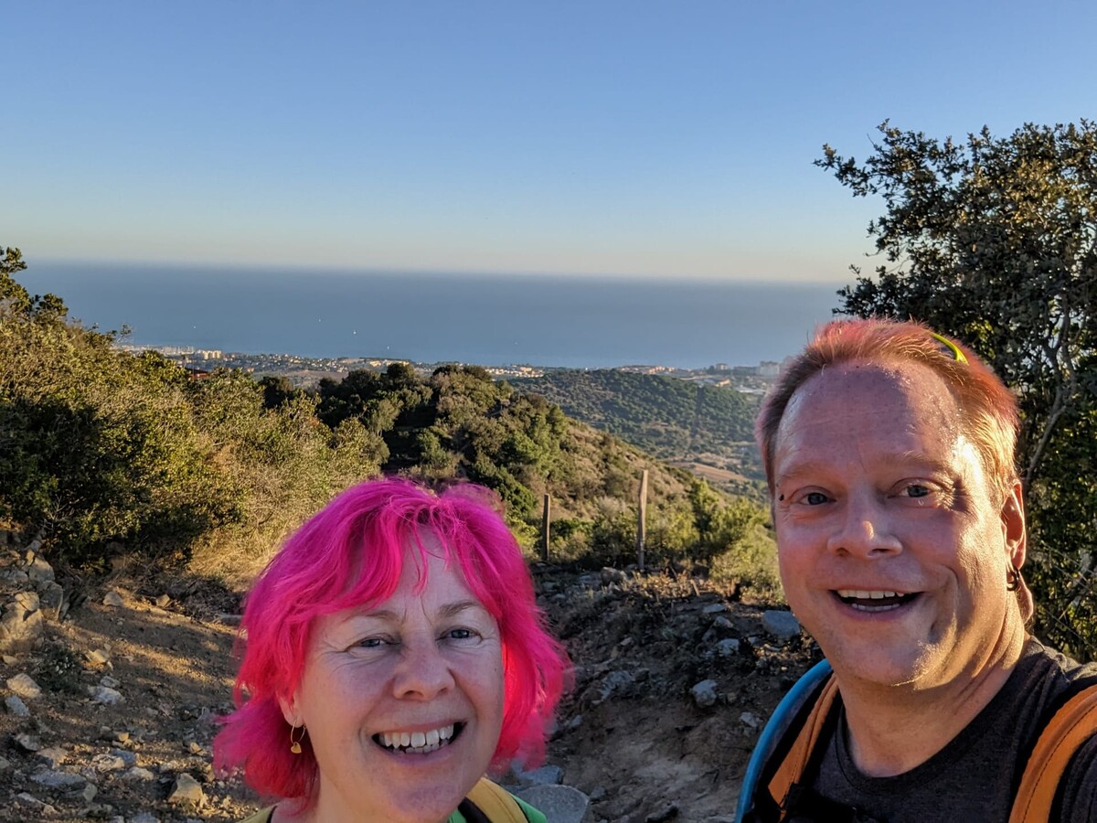 Selfie of Rob and Kirsten on a rocky hilltop trail, the Mediterranean Sea and a coastal town stretching behind them in warm golden hour light. Both grinning, Rob in a dark t-shirt with an orange backpack strap, Kirsten with bright magenta hair.