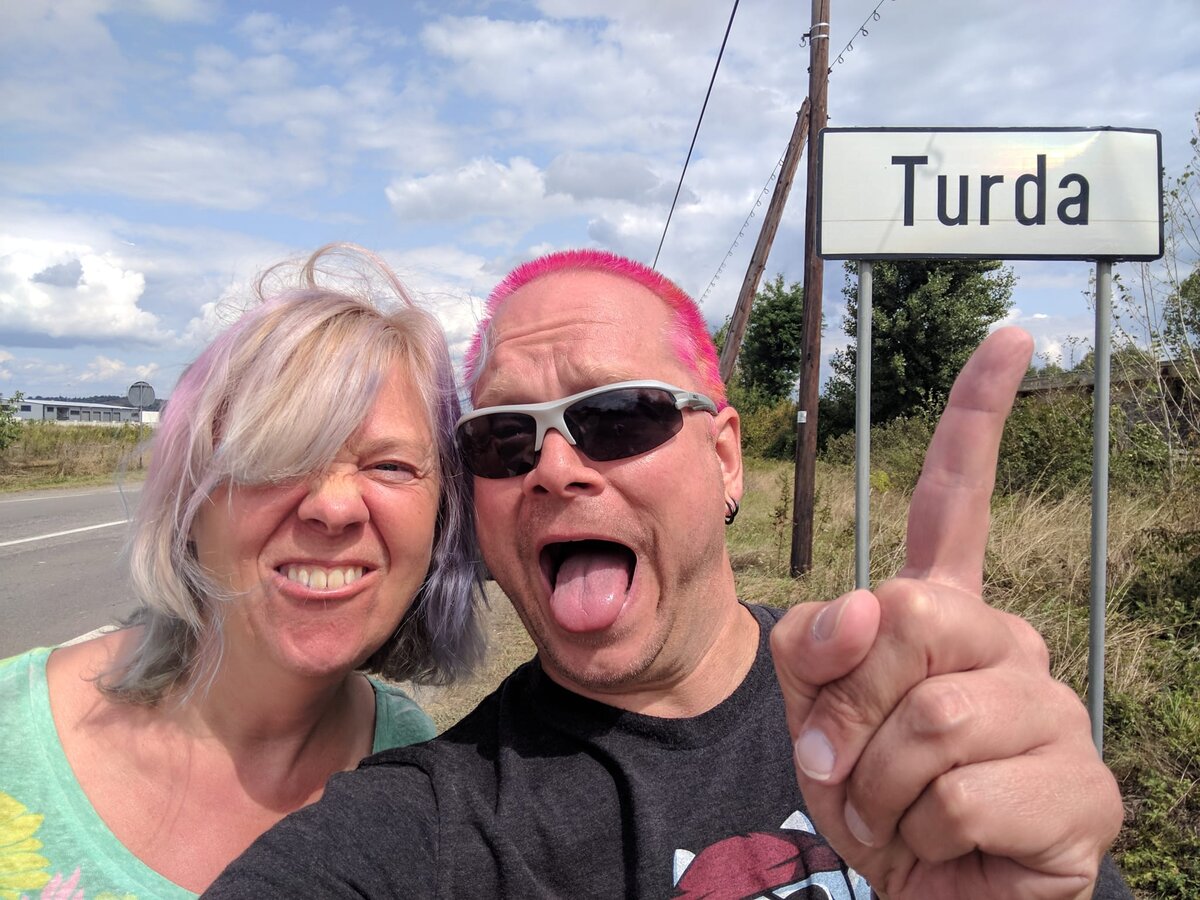 Rob with pink hair sticking his tongue out and Kirsten with silver-grey hair laughing beside him, the Turda road sign partially visible behind them on a rural Romanian roadside.
