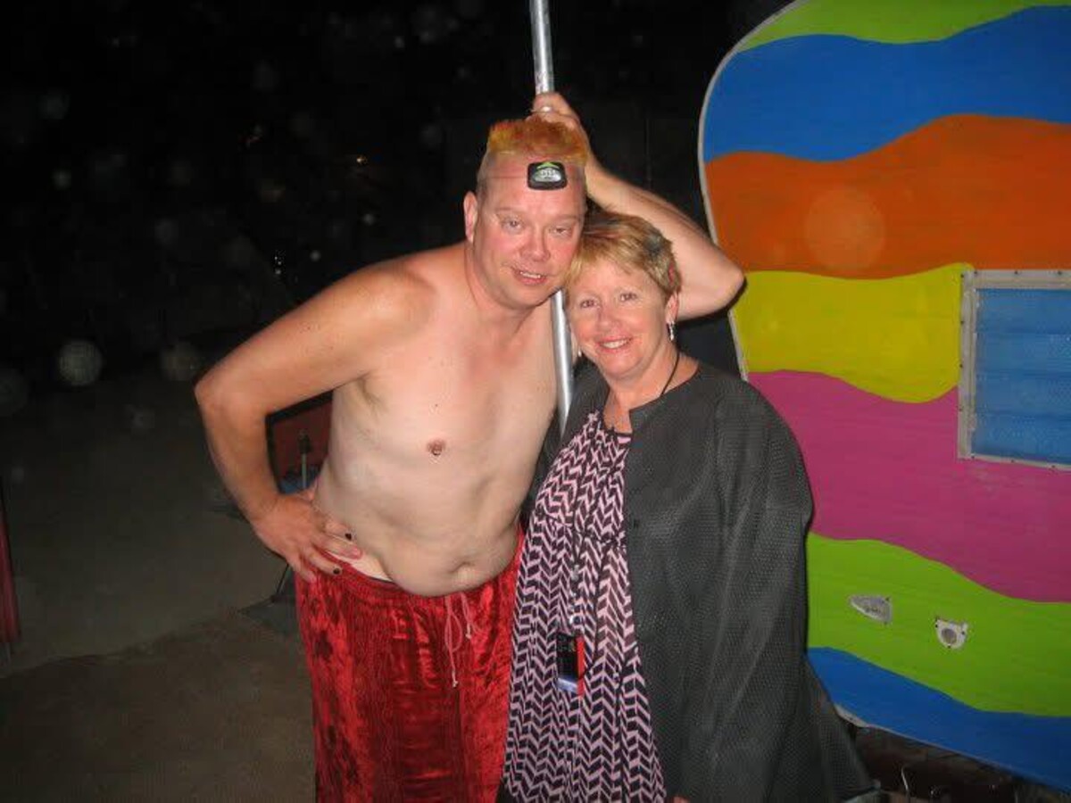 Rob shirtless with spiky orange-red hair and red crushed-velvet pants standing beside Lillith in a black chevron-print dress, a rainbow-striped trailer behind them on the Burning Man playa at night.