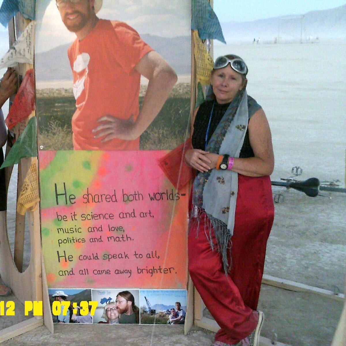 Lillith standing beside a memorial panel at Burning Man's Temple de Cortez, August 2012. The panel displays a large photo of Greg in an orange shirt and white hat, a poem about sharing both worlds of science and art, and smaller photos below. Lillith wears red pants, a grey scarf, and goggles pushed up on her head. The flat playa stretches behind under hazy sky.