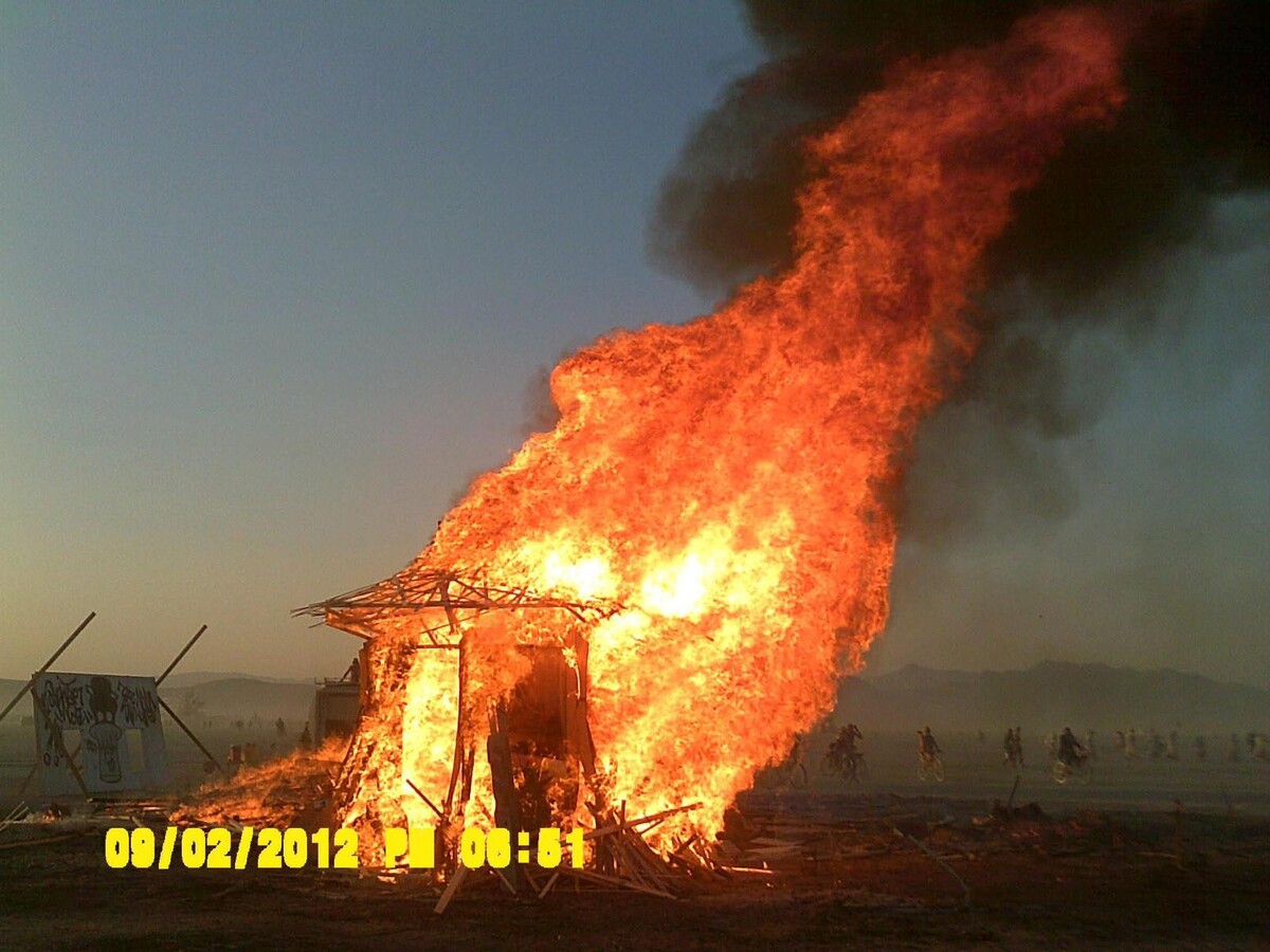 The Temple de Cortez engulfed in towering orange-yellow flames and black smoke on the Burning Man playa at twilight, the wooden structure collapsing, distant silhouettes of watchers on the horizon.