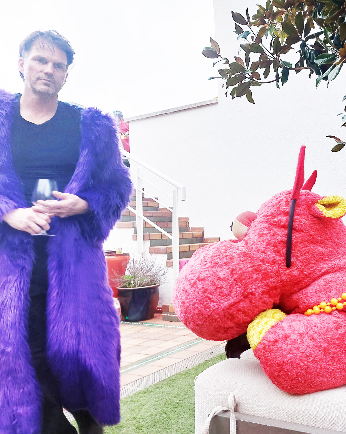 Large red stuffed hippo wearing a yellow beaded necklace, lounging on an outdoor seat at a garden gathering. A man in a full-length purple faux-fur coat stands beside it.