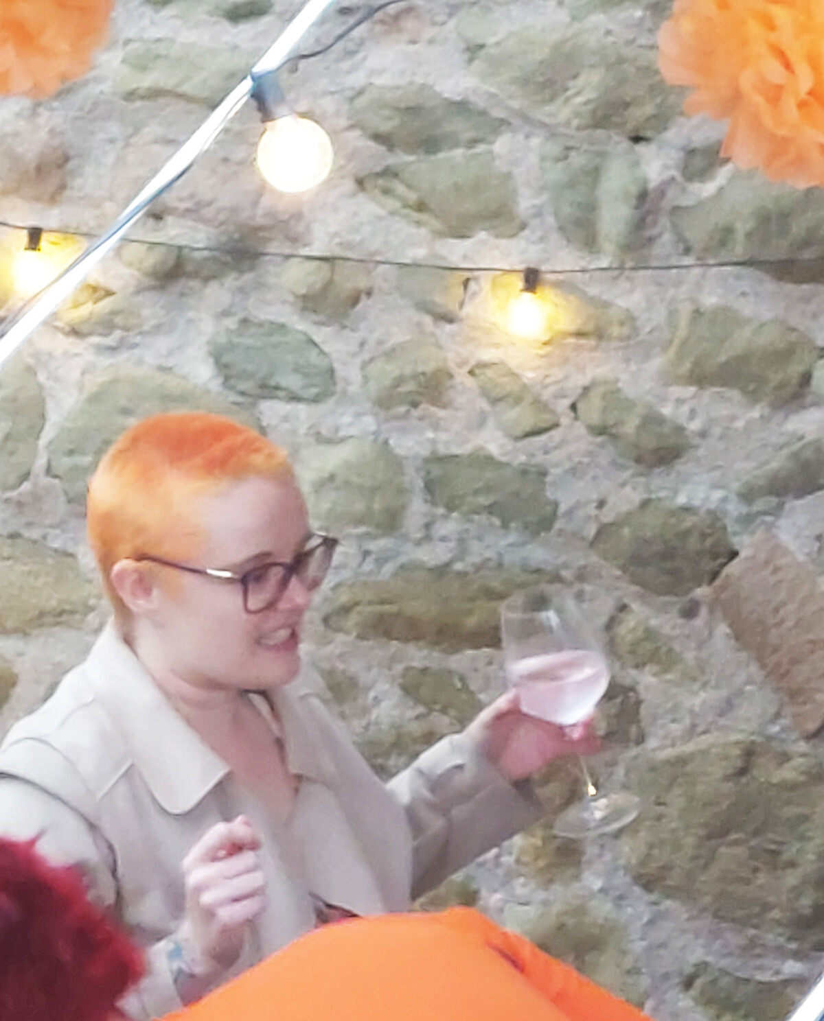 A woman with bright orange-red short hair raises a glass of ros&eacute; wine at an outdoor gathering decorated with orange paper pom-poms and string lights.