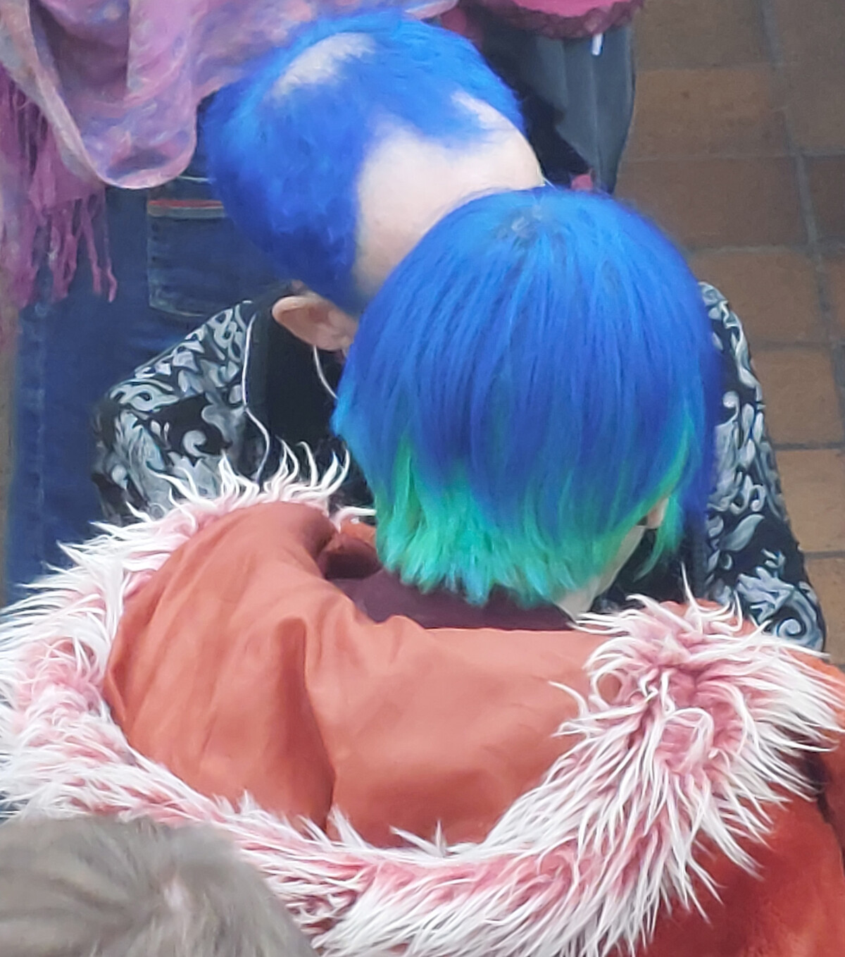 Two people with freshly dyed blue hair, foreheads touching, eyes closed, at an outdoor gathering.