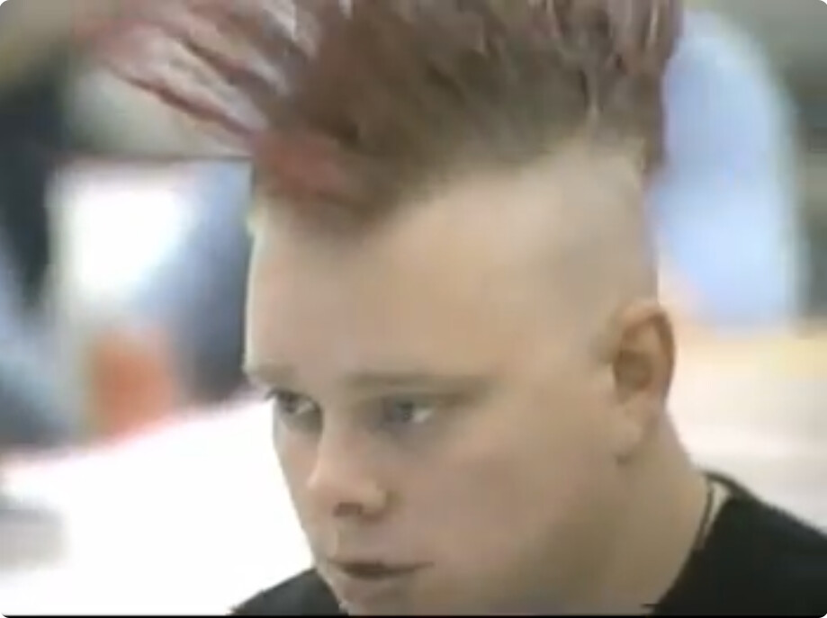 VHS video still of young Rob at the 1994 ACM programming contest finals, reddish-blond mohawk standing tall above shaved sides, black shirt, focused expression. Grainy broadcast-quality footage.