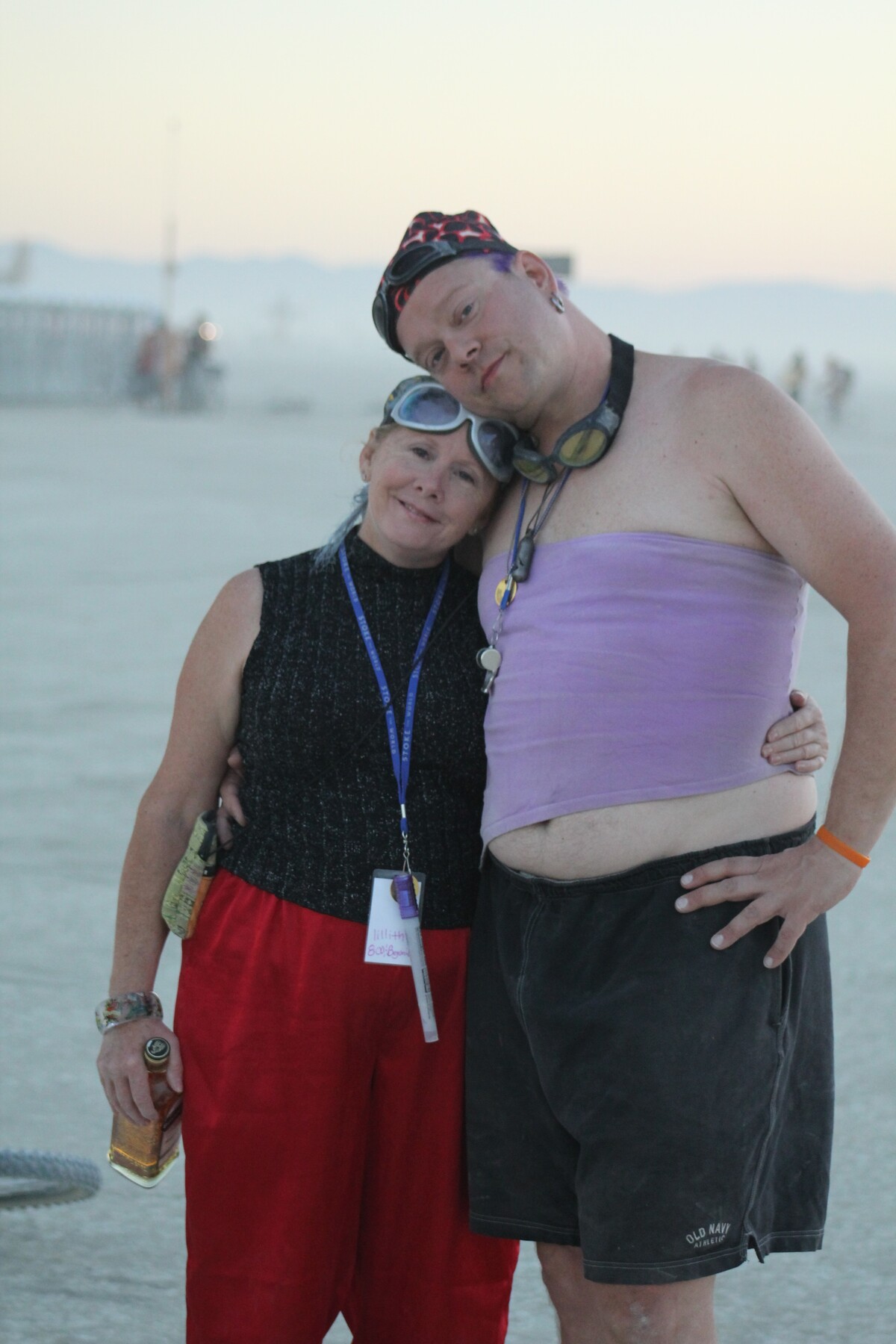 Rob and his adopted sister Lillith standing close on the Burning Man playa at dusk — Lillith's head tilted toward Rob's shoulder, soft pale sky behind them