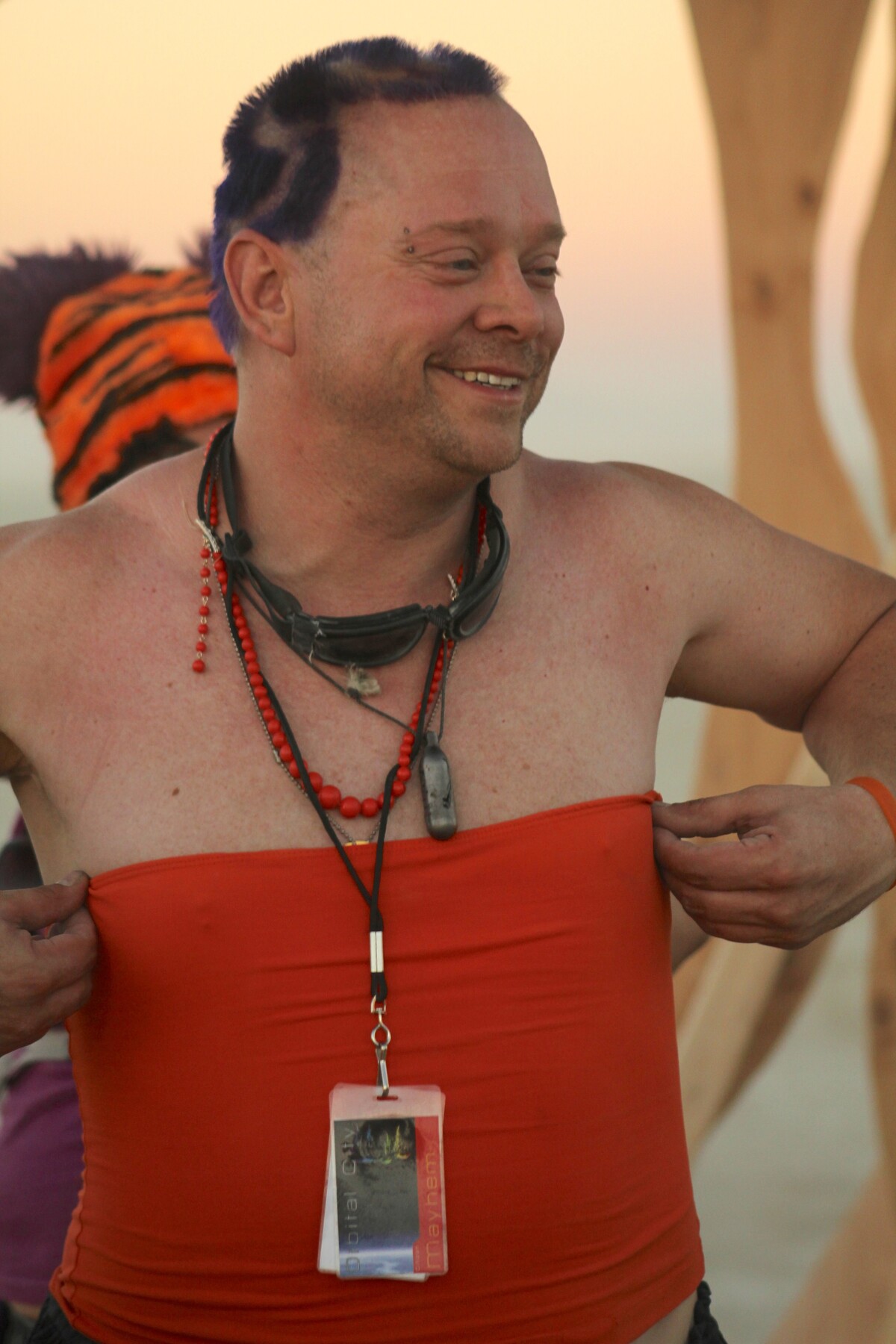 Rob grinning at Burning Man — blue-purple mohawk, red tube top, red beads, goggles around his neck, badge reading Mayhem