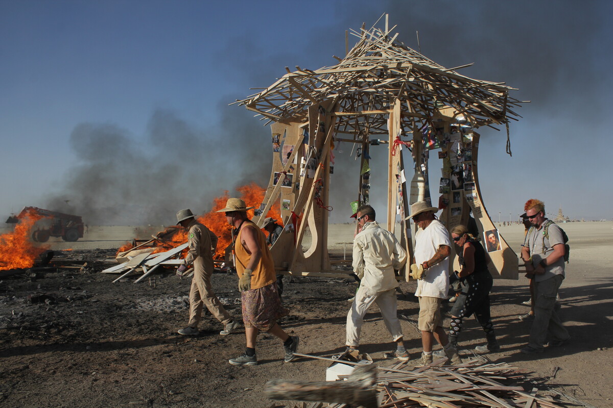 The Temple of Greg during the burn ceremony — flames rising behind it, crew members walking through the burn site, the structure still standing