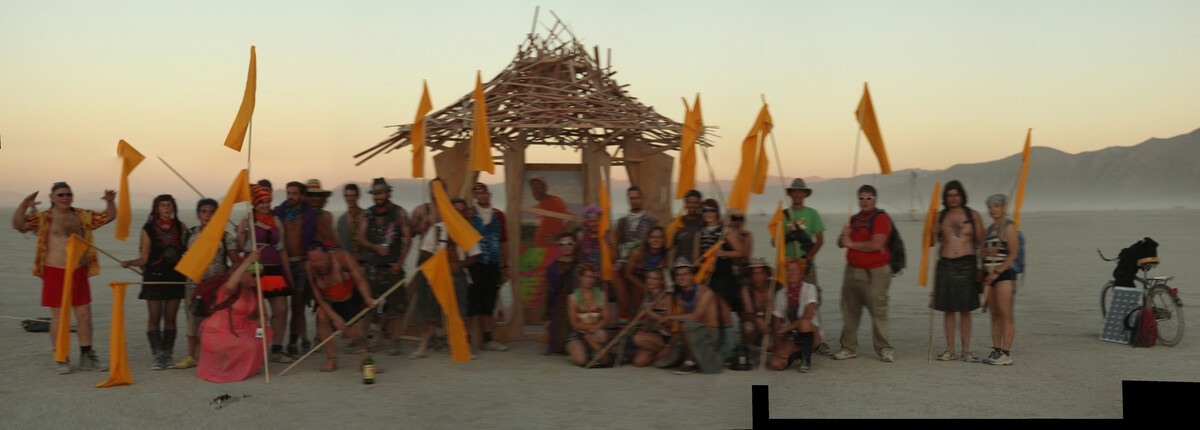 Panoramic group photo of 30+ people holding yellow flags in front of the Temple of Greg at sunset on the Burning Man playa