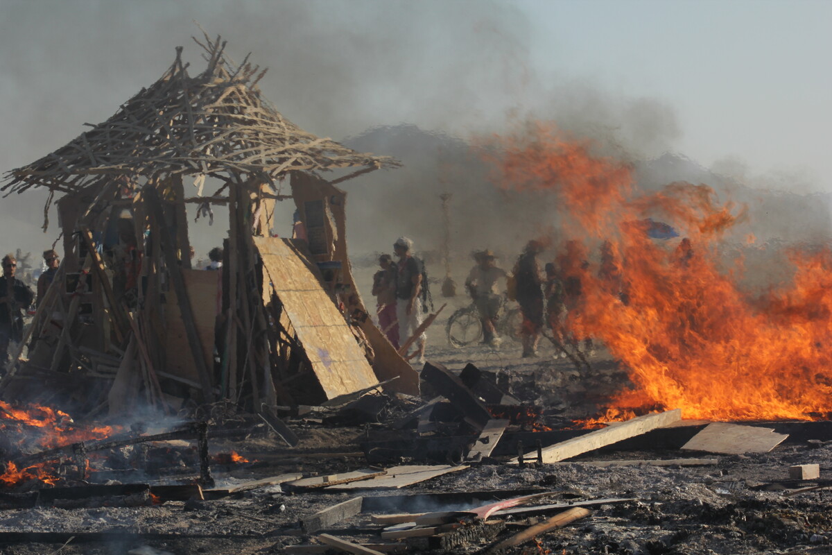 The Temple of Greg mid-collapse, engulfed in massive flames, silhouettes of watchers visible through smoke on the Burning Man playa
