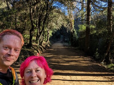Selfie of Rob and Kirsten on a sunlit forest trail, a long dirt path stretching behind them through gnarled moss-covered trees forming a natural tunnel